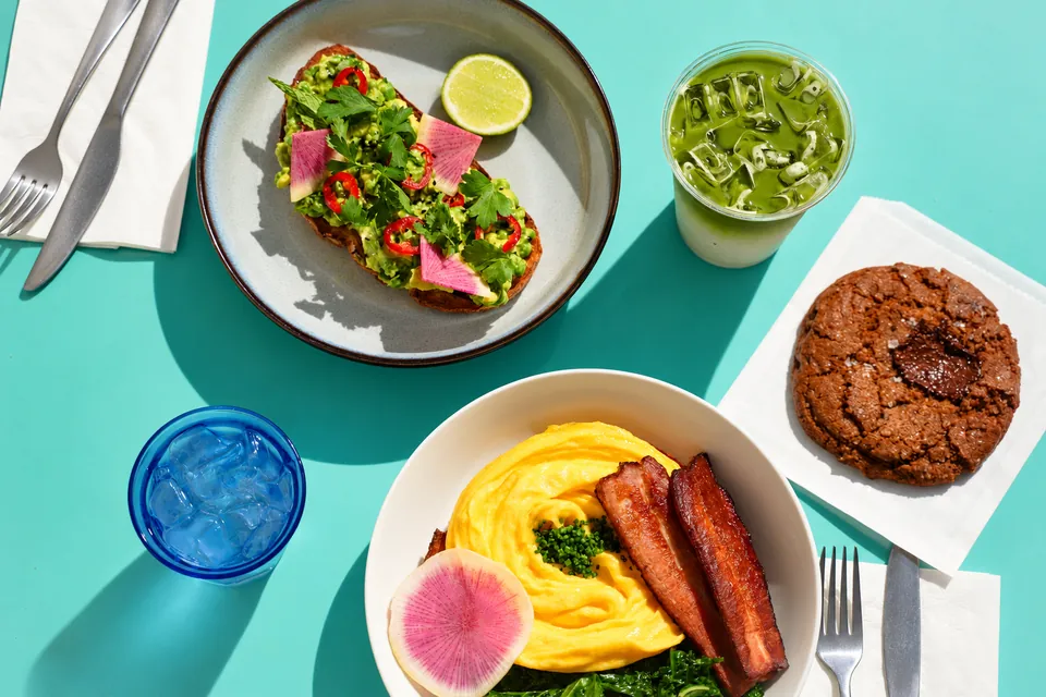 Avocado toast, bacon and eggs, matcha and a cookie on a sunny turquoise café table
